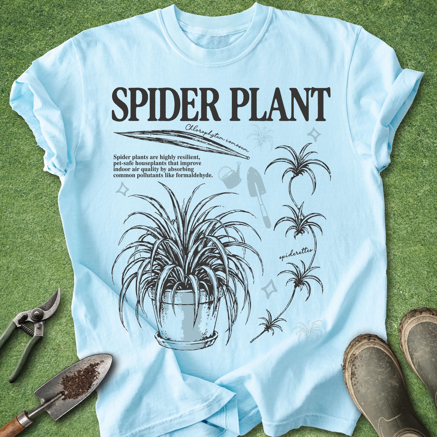 A light blue graphic t-shirt laid flat on green grass, featuring black text that reads 'SPIDER PLANT' with botanical illustrations of a spider plant. Gardening props including pruning shears, a trowel, and boots are visible around the shirt.