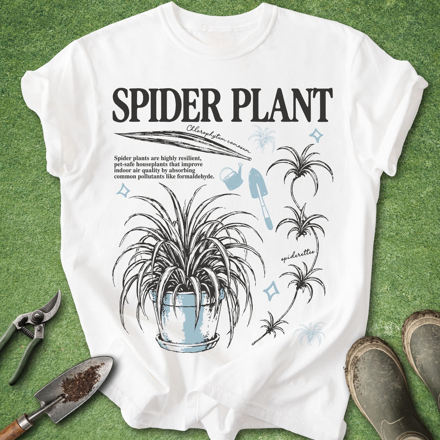A white graphic T-shirt laid flat on green grass-like ground, with gardening tools and boots around it. The shirt features large black text reading 'SPIDER PLANT' and a black-and-light-blue spider plant illustration with small decorative text.