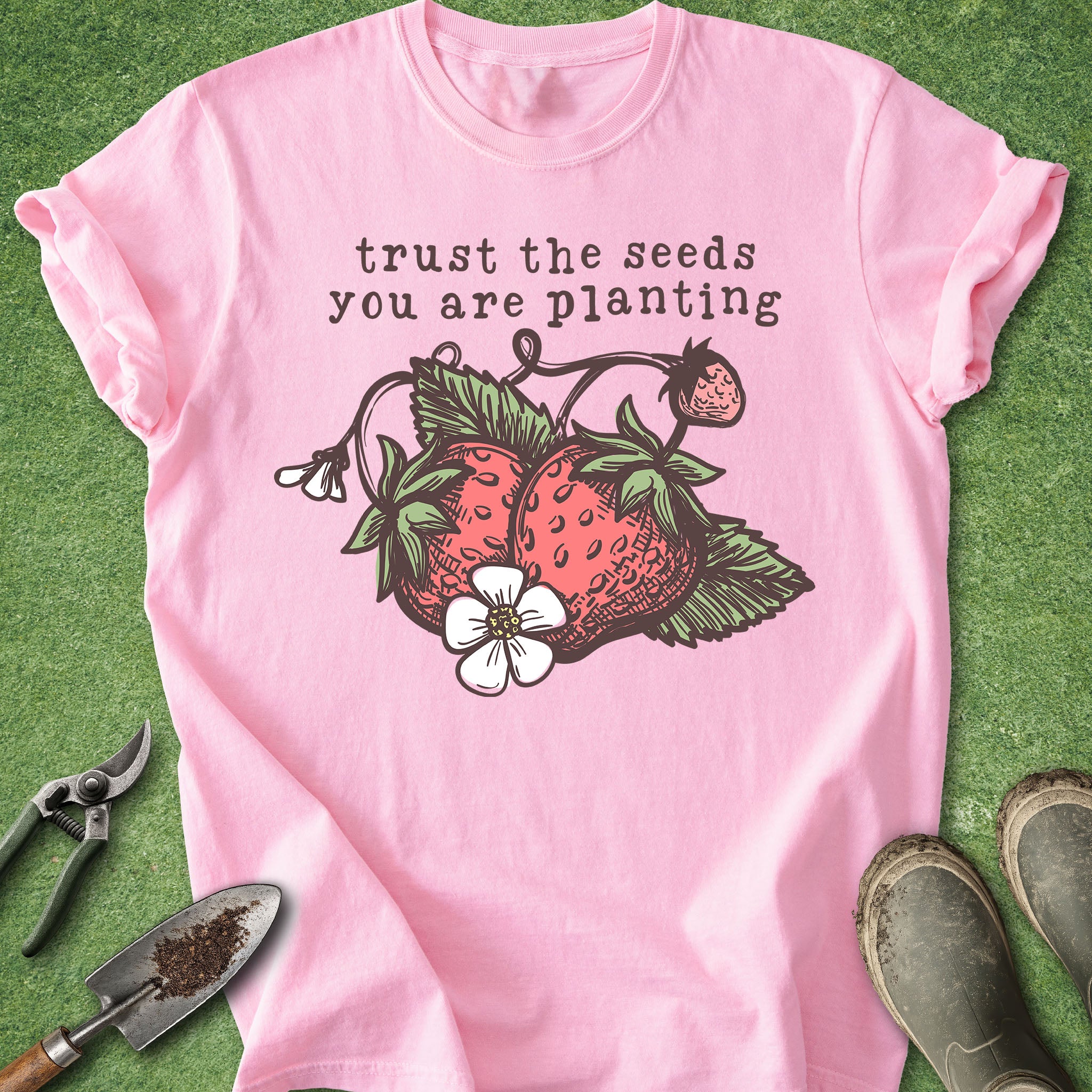 Pink t-shirt with strawberry graphic and text 'trust the seeds you are planting' on a green background