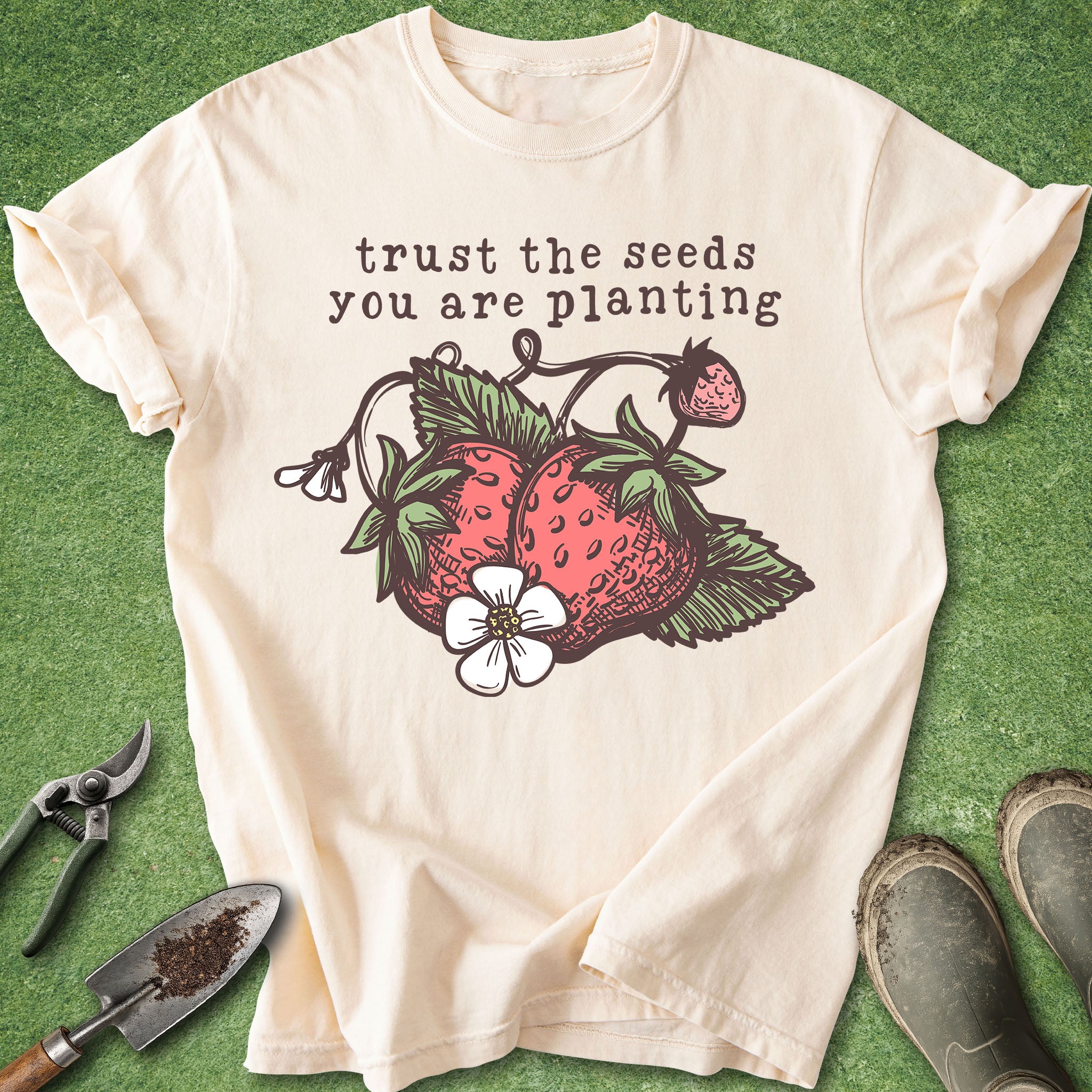 Beige t-shirt with strawberry graphic and text 'trust the seeds you are planting' on a green background