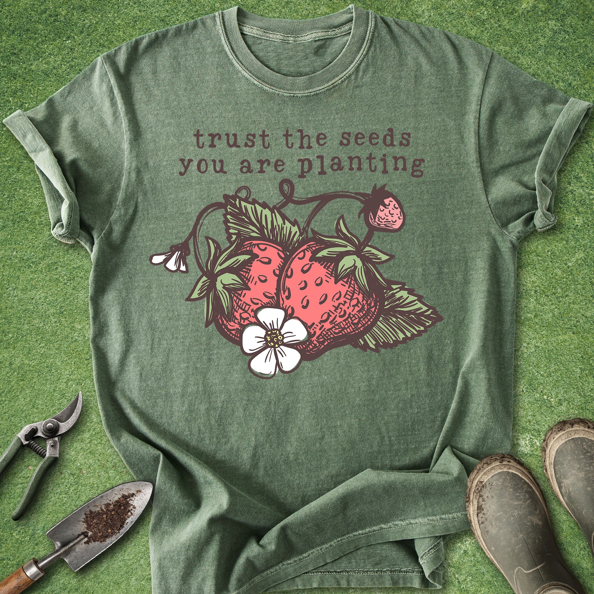 Green t-shirt with strawberry design and text 'trust the seeds you are planting' on a grass background.