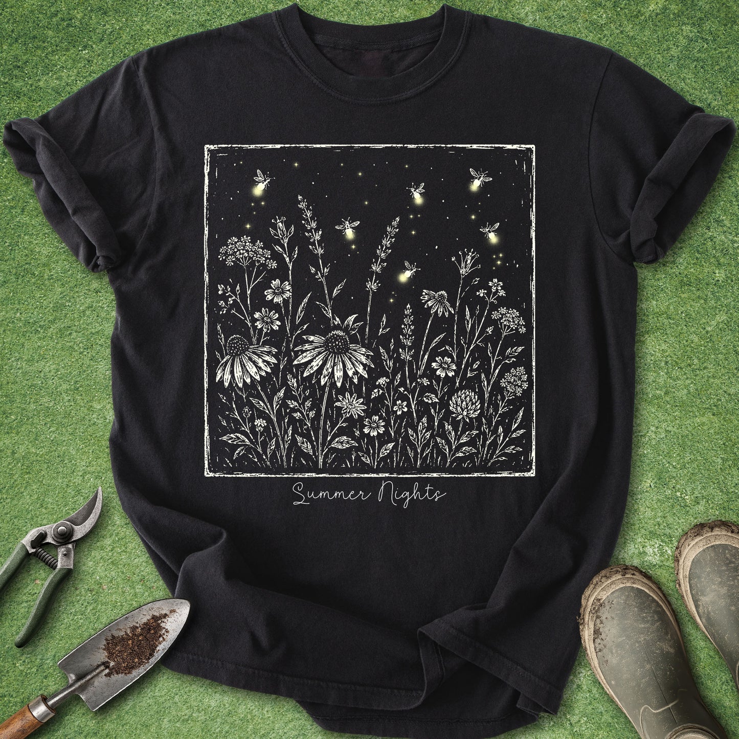 A black short-sleeve graphic T-shirt laid flat on green grass, featuring a white floral and firefly design with the words 'Summer Nights.' Gardening tools and boots are partially visible around the shirt as styling props.