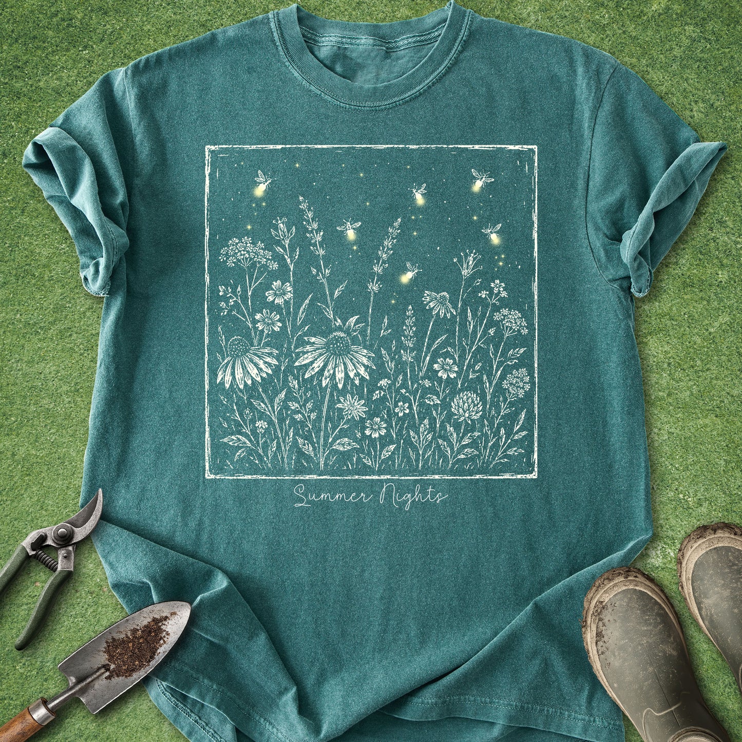 A teal short-sleeve t-shirt laid flat on green grass, featuring a white square graphic of wildflowers and glowing fireflies with the text 'Summer Nights' below. Gardening tools and muddy boots are placed around the shirt as props.