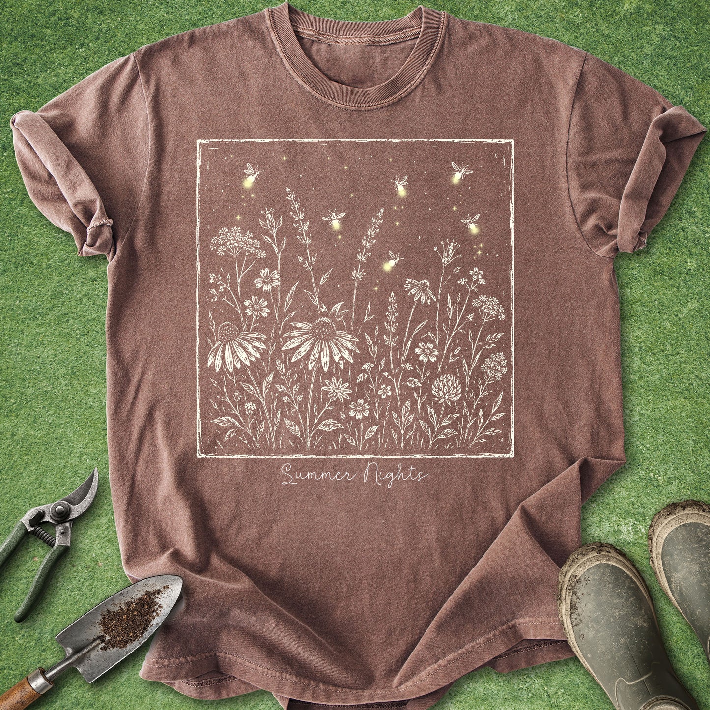 A brown short-sleeve t-shirt with a white and yellow 'Summer Nights' firefly and wildflower graphic, laid flat on green grass with gardening tools and boots nearby.