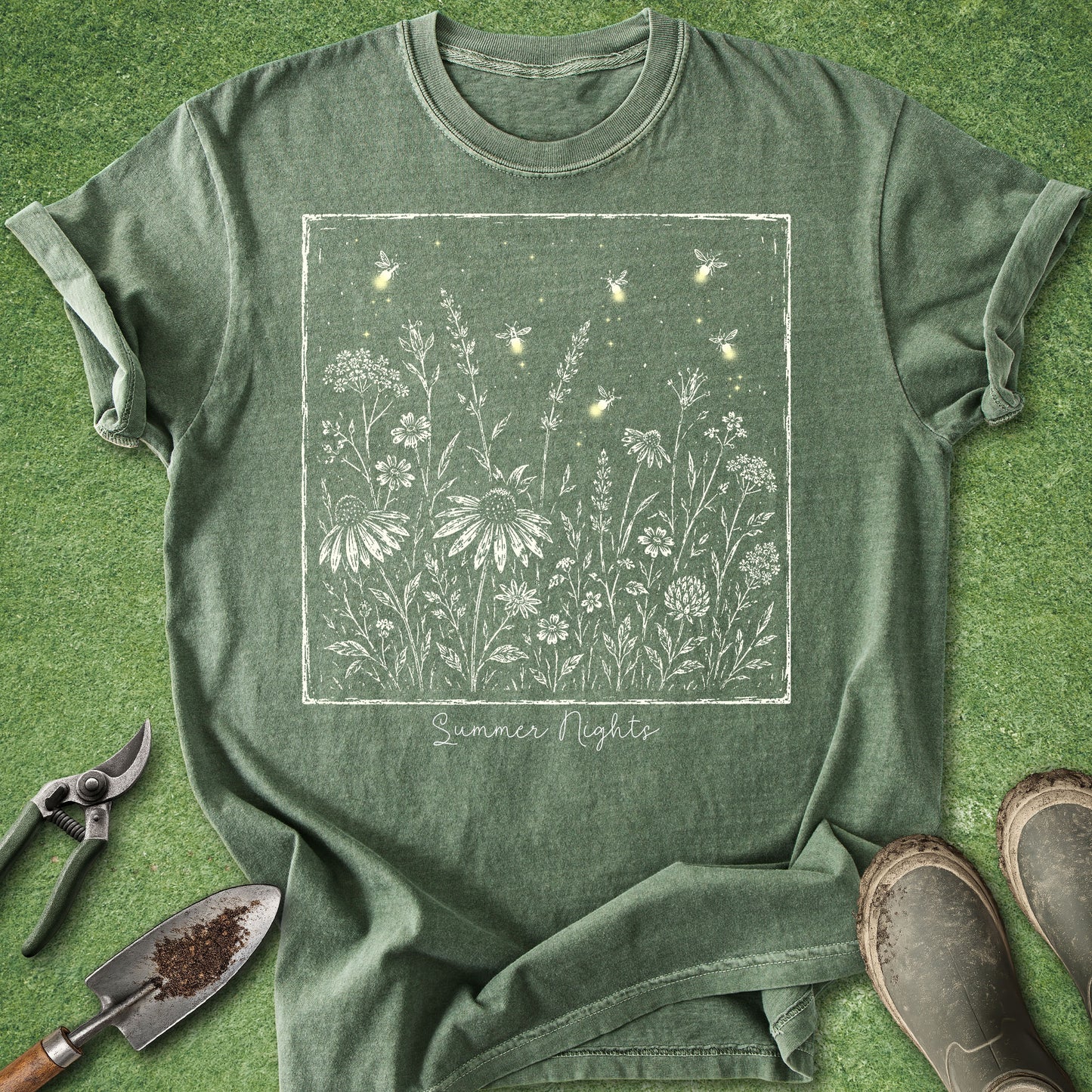 An olive green short-sleeve t-shirt with a white line-art graphic of wildflowers, fireflies, and stars, with the text 'Summer Nights' printed below. The t-shirt is laid flat on a green grass-like surface, styled with gardening tools (trowel, pruning shears) and work boots.