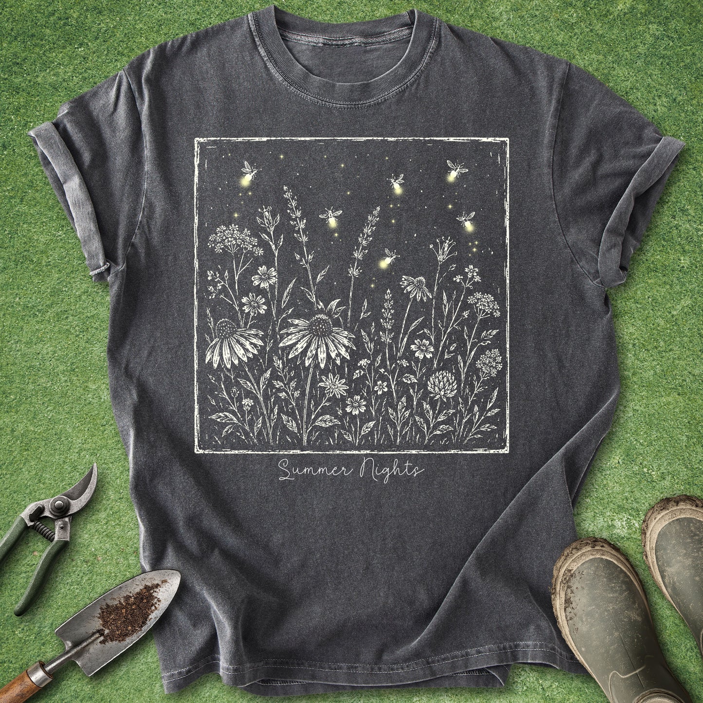 A dark charcoal gray graphic t-shirt laid flat on green grass, surrounded by gardening props including pruning shears, a trowel, and work boots. The t-shirt features a square graphic of white wildflowers and glowing fireflies on a dark background, with the text 'Summer Nights' written below.