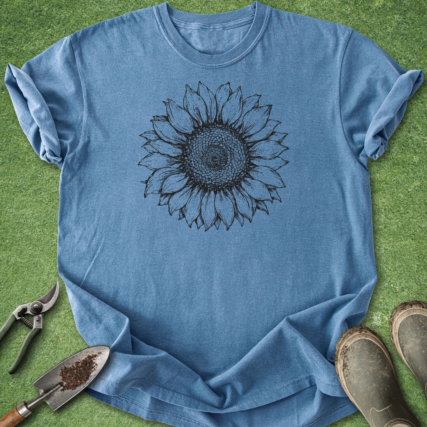 A light blue crewneck t-shirt with a black sunflower graphic printed on the chest, laid flat on green grass with gardening tools and boots around it.