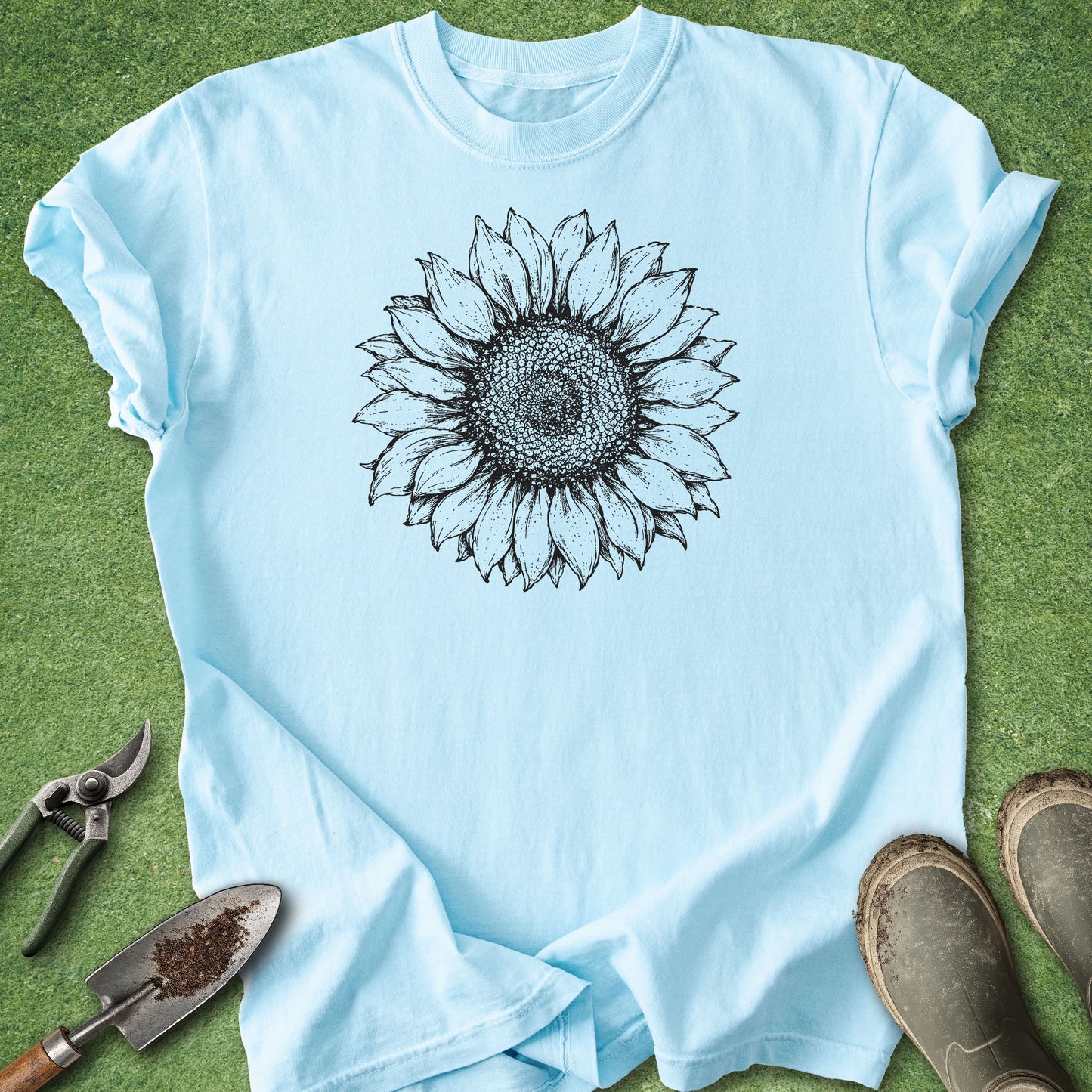 A light blue short-sleeve t-shirt with a black line art sunflower graphic on the chest, laid flat on green grass with gardening tools and boots nearby.