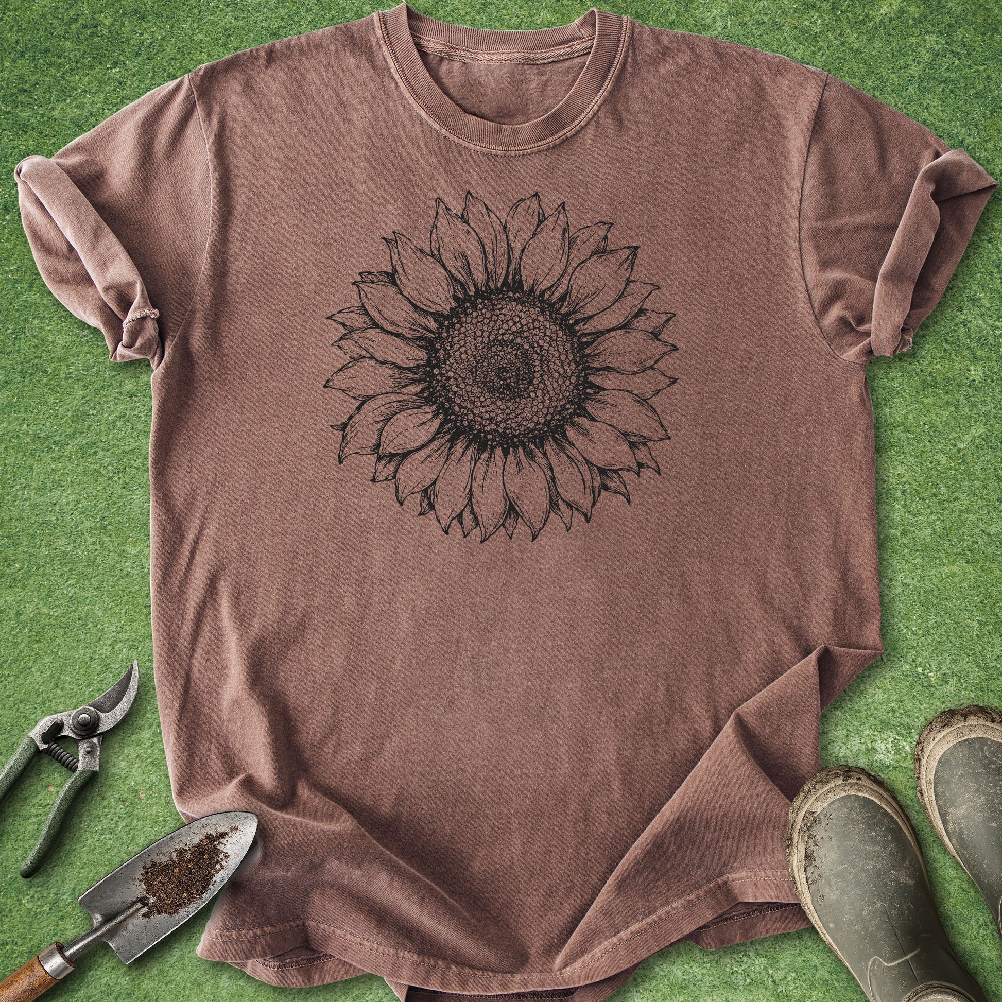 A brown short-sleeve t-shirt with a black sunflower graphic printed on the chest, laid flat on green grass with gardening props including pruning shears, a trowel, and boots.