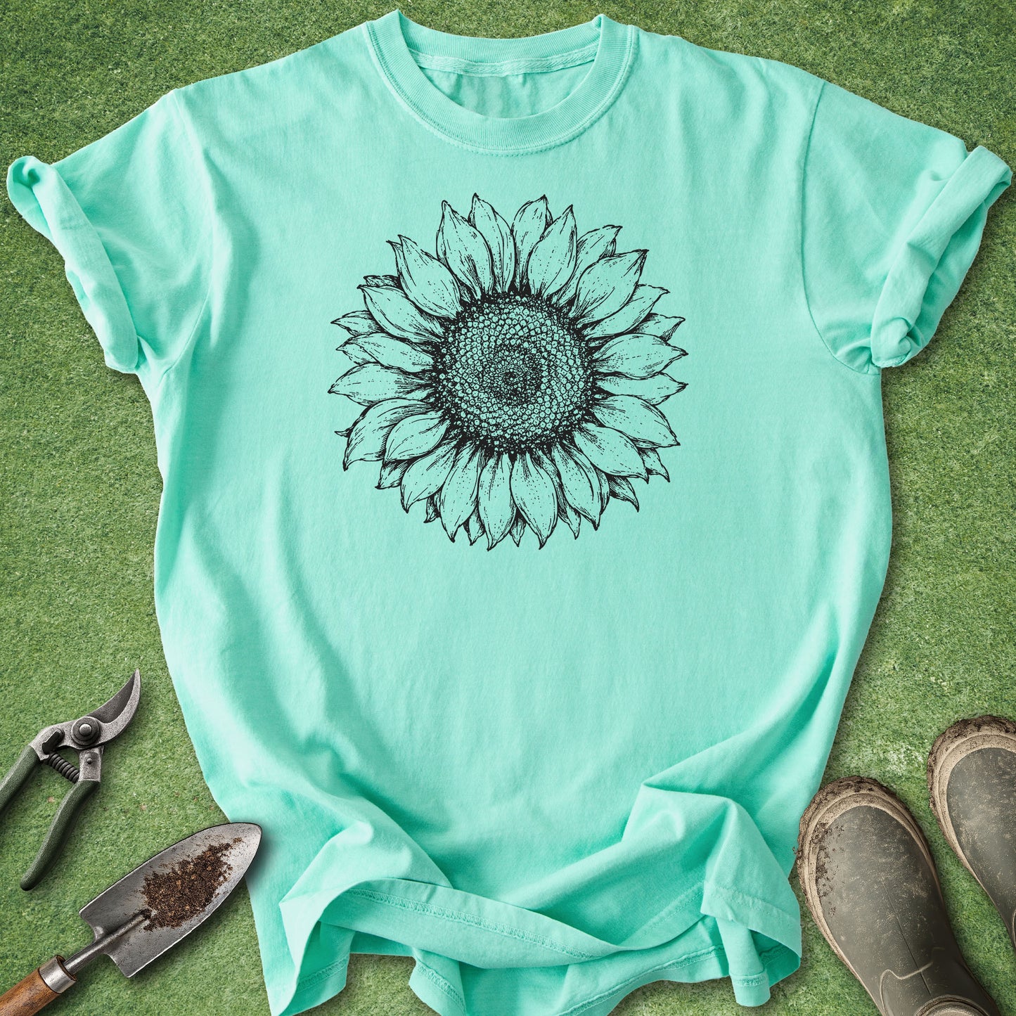 A mint green short-sleeve t-shirt with a black sunflower graphic printed on the chest, laid flat on green grass with gardening tools and boots nearby.