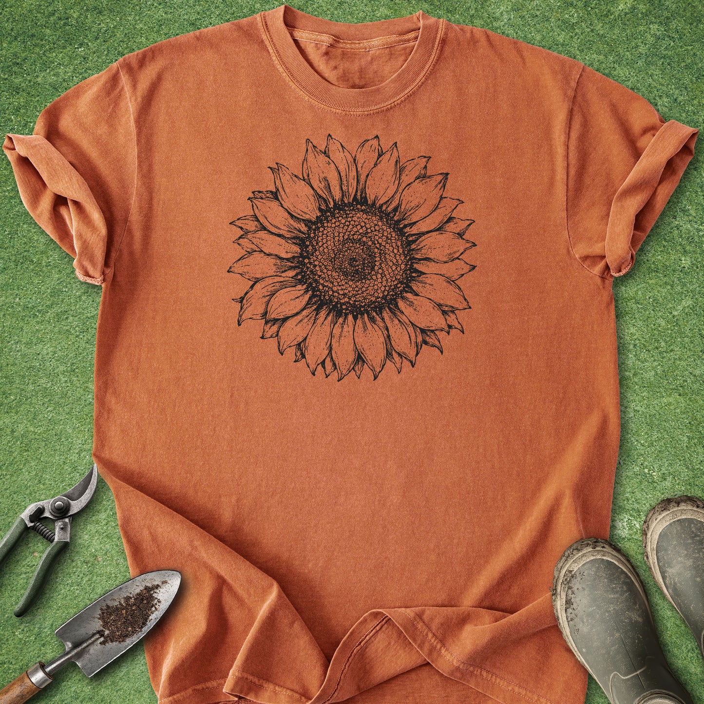 An orange short-sleeve t-shirt with a black sunflower graphic printed on the chest, laid flat on green grass with gardening tools and boots around it.