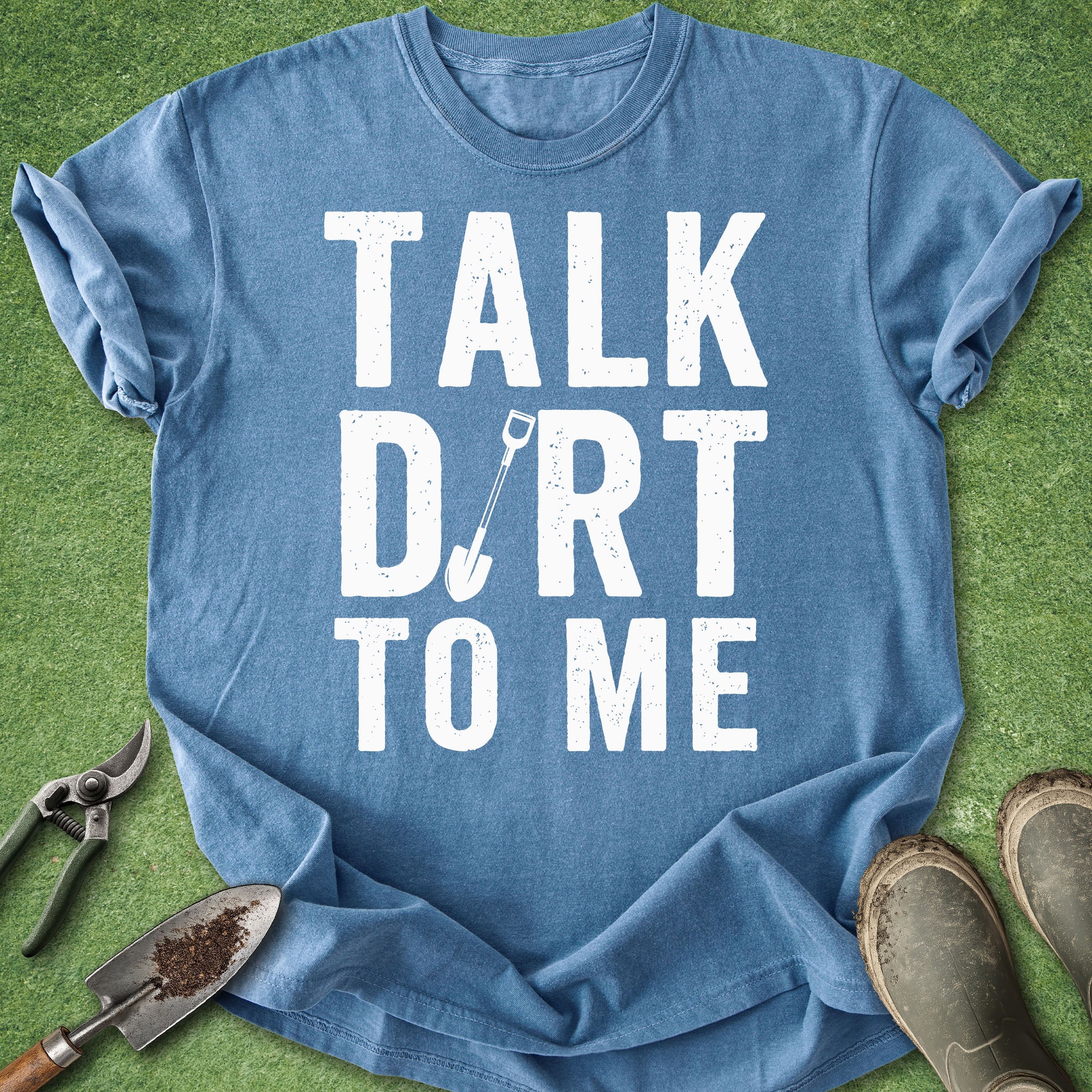 Blue t-shirt with 'Talk Dirt to Me' text on a grass background with gardening tools.
