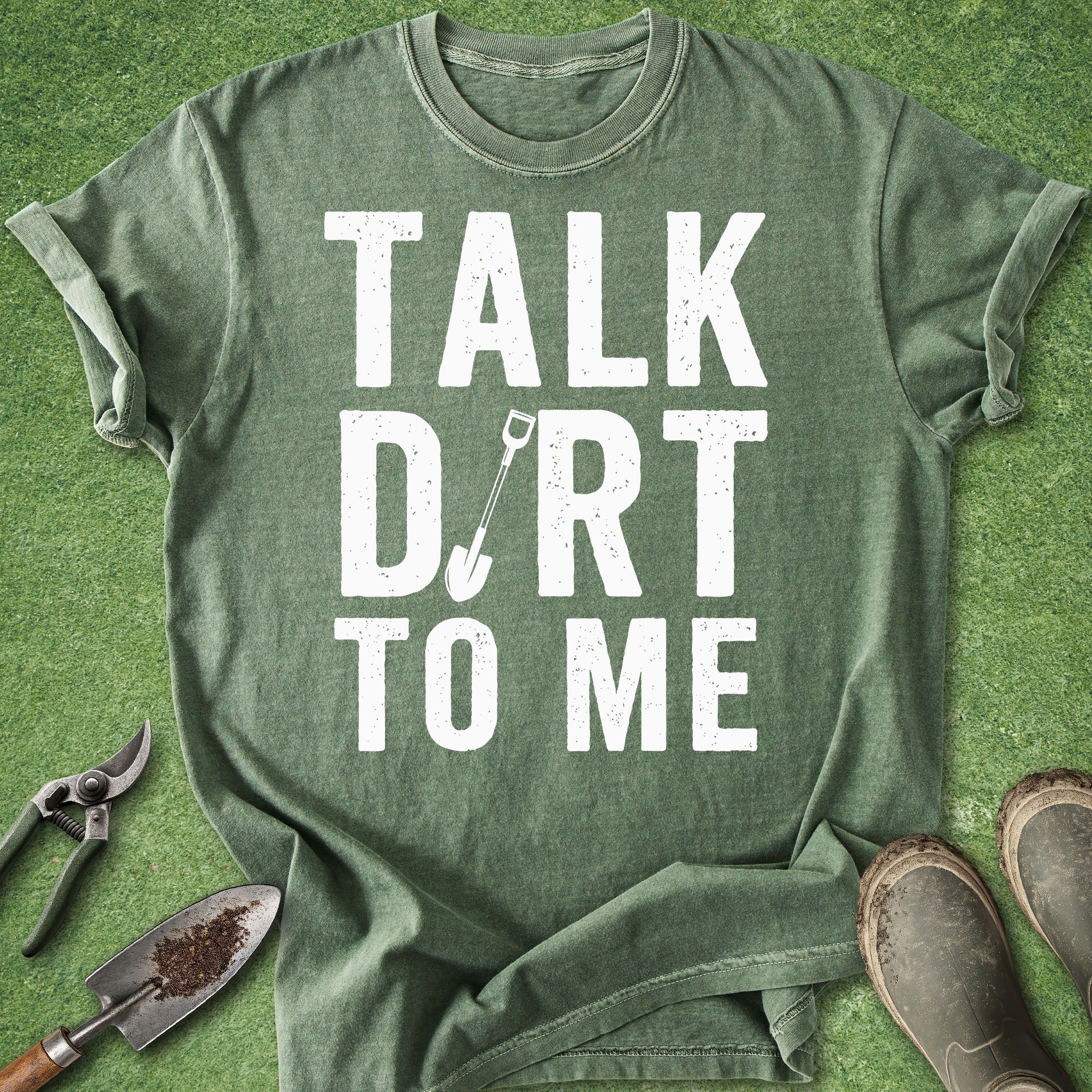 Green t-shirt with 'Talk Dirt to Me' text on a grass background with gardening tools.