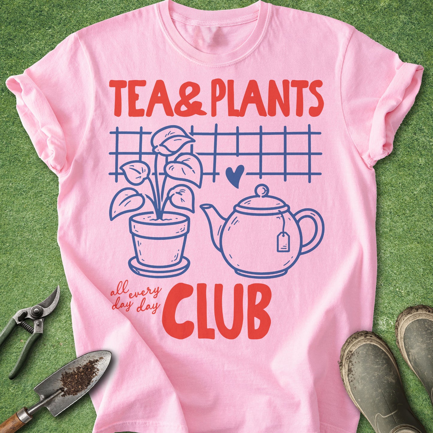 A pink short-sleeve graphic T-shirt laid flat on green grass-like ground, with gardening tools and boots around it. The shirt reads 'TEA & PLANTS' and 'all every day CLUB' in red text, with a blue teapot, potted plant, heart, and grid design on the front.