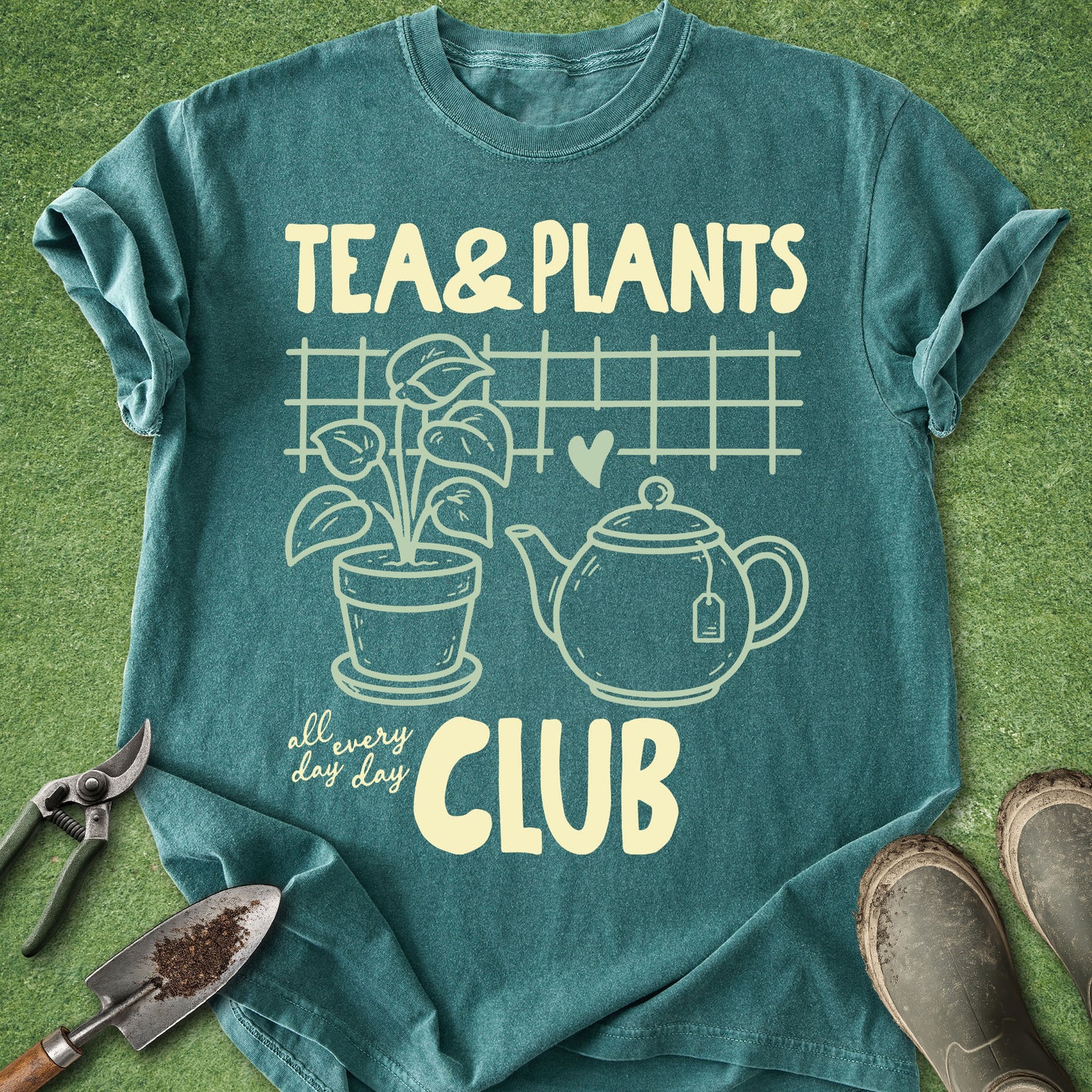 A teal graphic t-shirt laid flat on a green grass-like surface, featuring cream-colored text and illustrations that read 'TEA & PLANTS all every day CLUB' with a plant, teapot, and heart. Gardening props including pruning shears, a small hand trowel, and boots are placed around the shirt.
