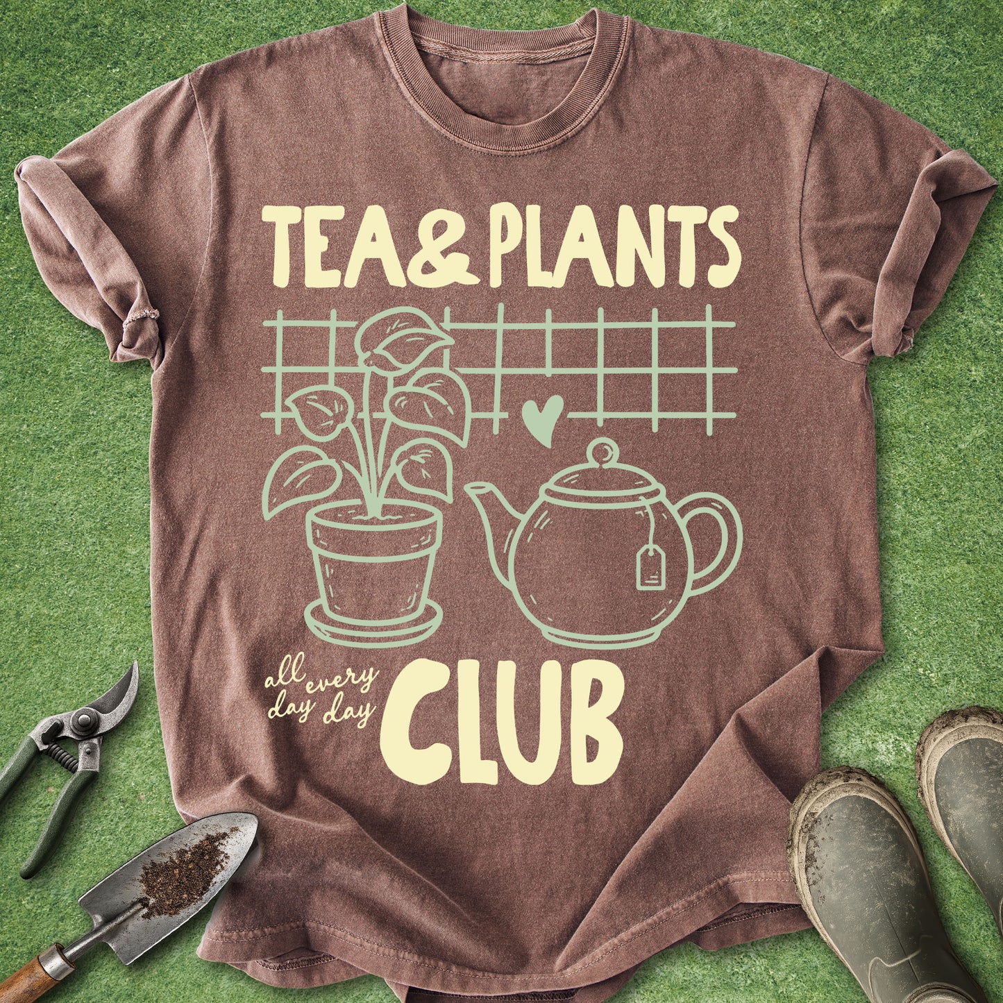 A brown short-sleeve graphic T-shirt laid flat on green grass-like ground, with gardening tools and boots around it. The shirt reads “TEA & PLANTS CLUB” in pale yellow text with a plant, teapot, heart, and grid illustration on the front.