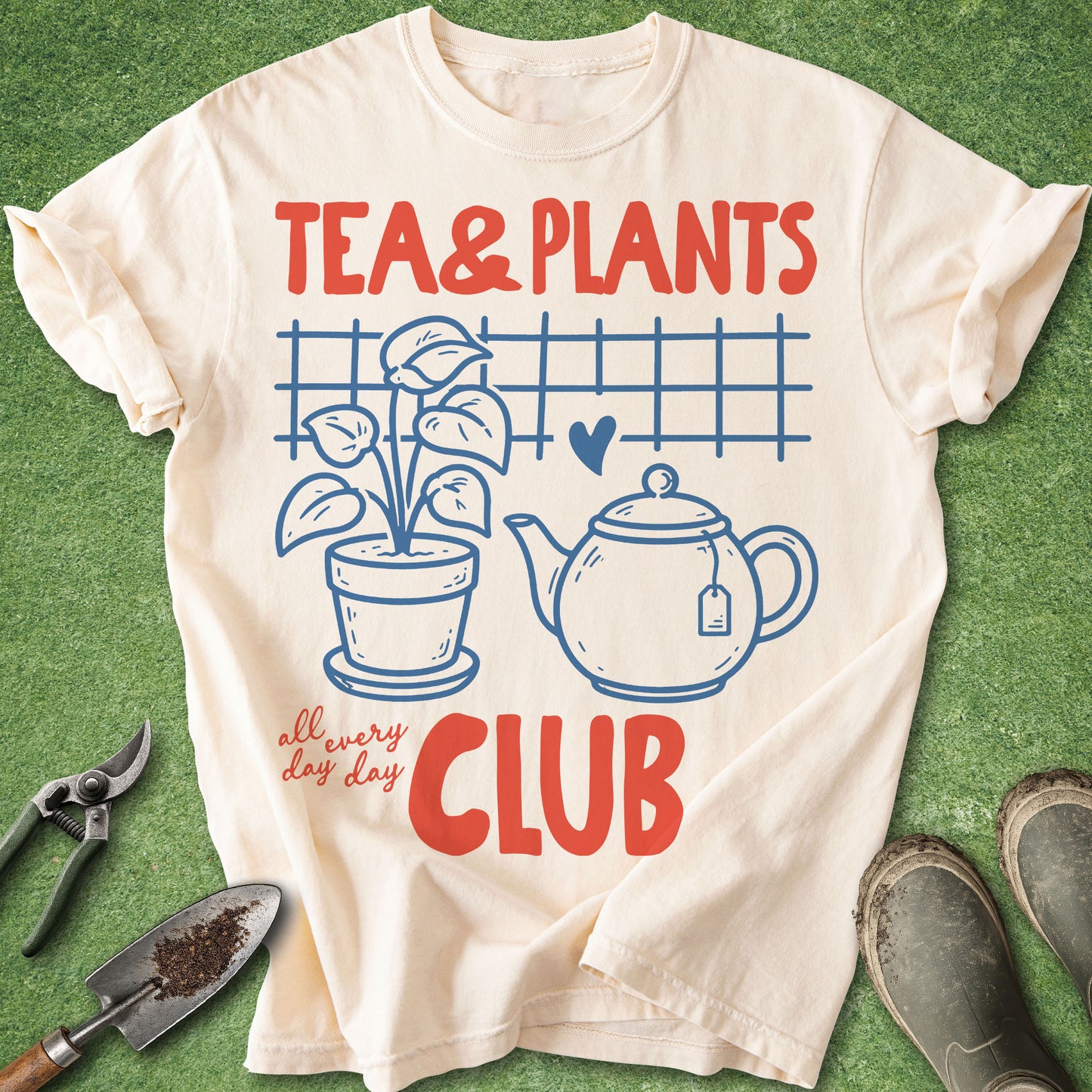 An off-white graphic t-shirt laid flat on green grass, featuring red and blue illustrations of a potted plant, teapot, heart, and the text 'TEA & PLANTS all every day CLUB'. Gardening tools and muddy boots are placed around the shirt as props.