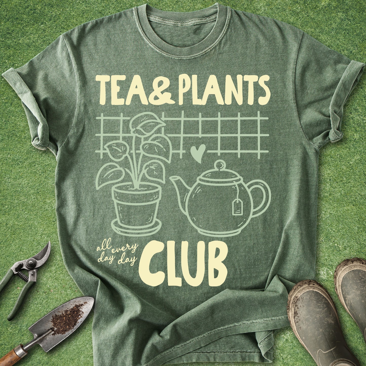 An olive green graphic t-shirt laid flat on a green grass-like surface, featuring cream-colored text and illustrations that read 'TEA & PLANTS' and 'all every day CLUB' with a teapot, plant, and fence design. Gardening props including pruning shears, a trowel, and boots are placed around the shirt.