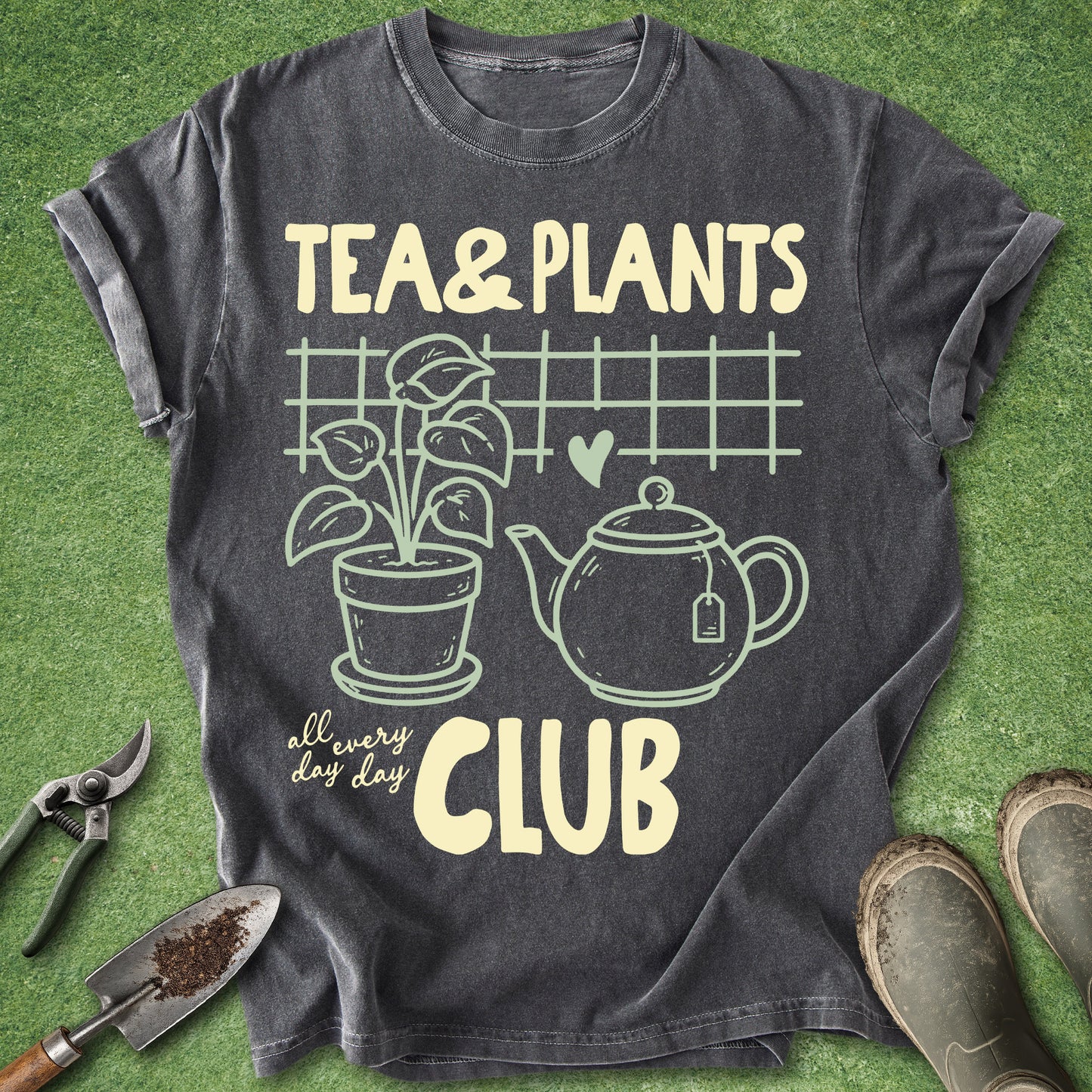 A dark charcoal gray graphic t-shirt laid flat on green grass, featuring a cream-colored print that reads 'TEA & PLANTS all every day CLUB' with illustrations of a potted plant, teapot, and grid. Gardening props including pruning shears, a trowel, and boots are arranged around the shirt.