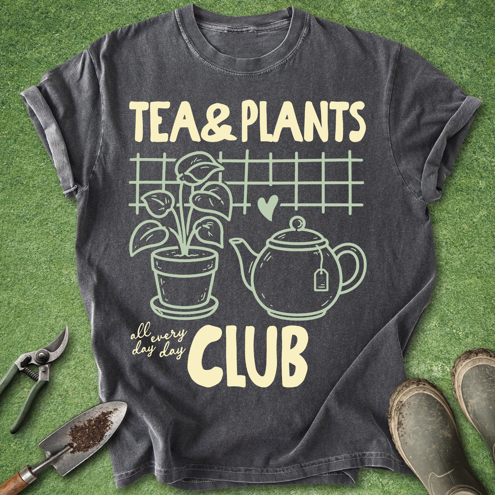 A dark charcoal gray graphic t-shirt laid flat on green grass, featuring a cream-colored print that reads 'TEA & PLANTS all every day CLUB' with illustrations of a potted plant, teapot, and grid. Gardening props including pruning shears, a trowel, and boots are arranged around the shirt.