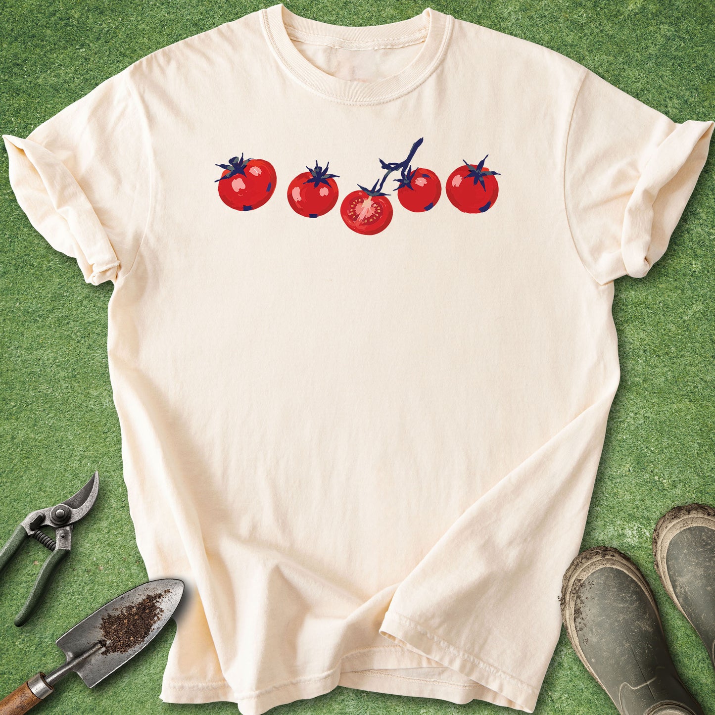 Beige t-shirt with tomato design on a green surface with gardening tools.