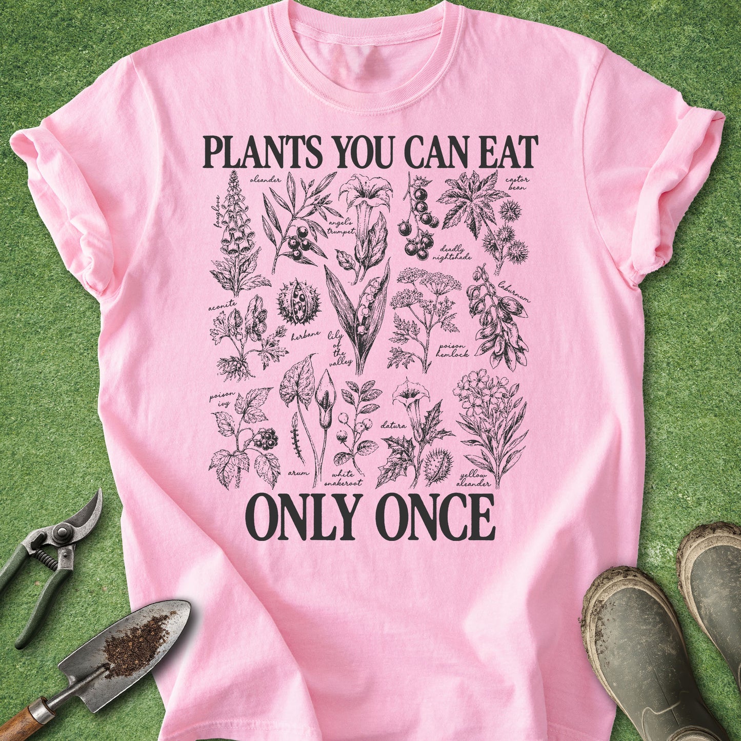 A pink short-sleeve graphic T-shirt laid flat on green grass-like ground, with gardening tools and boots around it. The shirt has black text reading 'PLANTS YOU CAN EAT ONLY ONCE' with a central botanical illustration print.