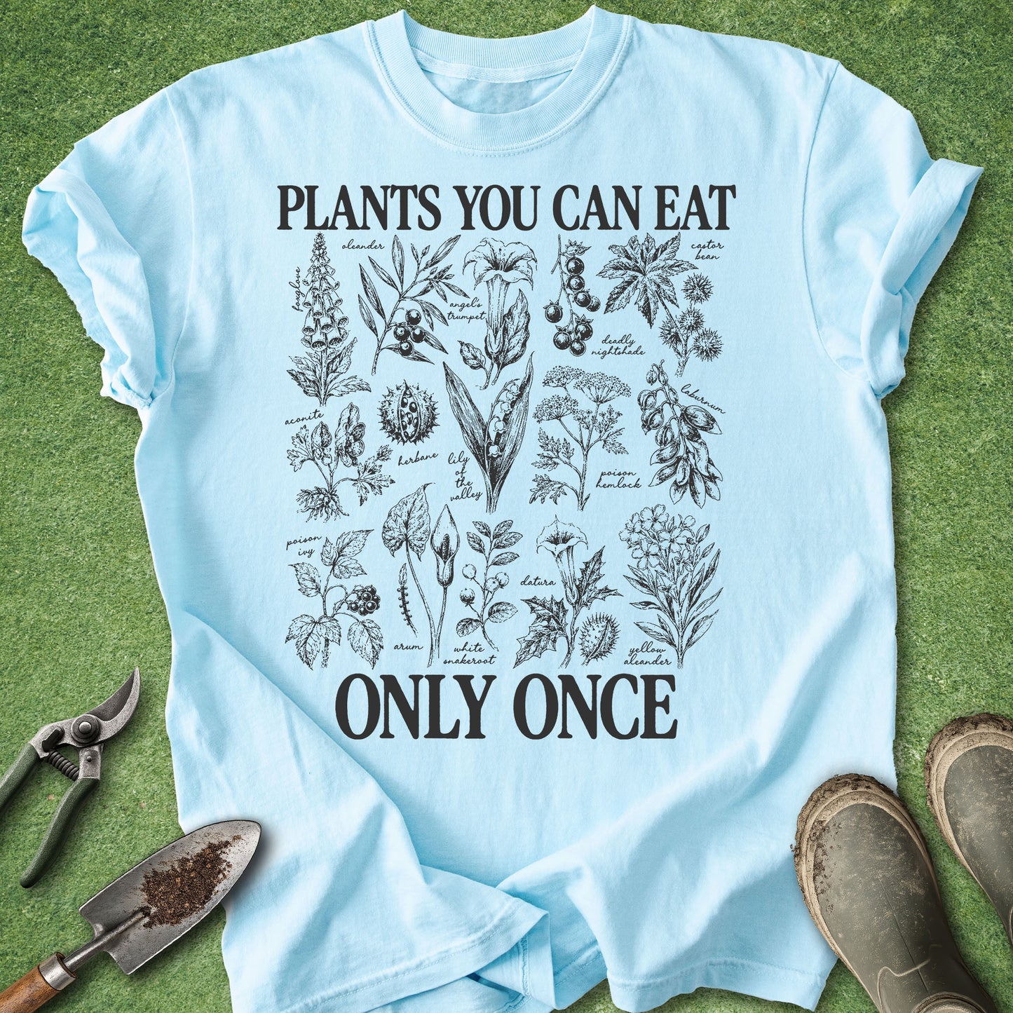 A light blue graphic t-shirt laid flat on green grass-like ground, with gardening tools and boots around it. The shirt has black text reading 'PLANTS YOU CAN EAT ONLY ONCE' above and below a detailed botanical illustration print.