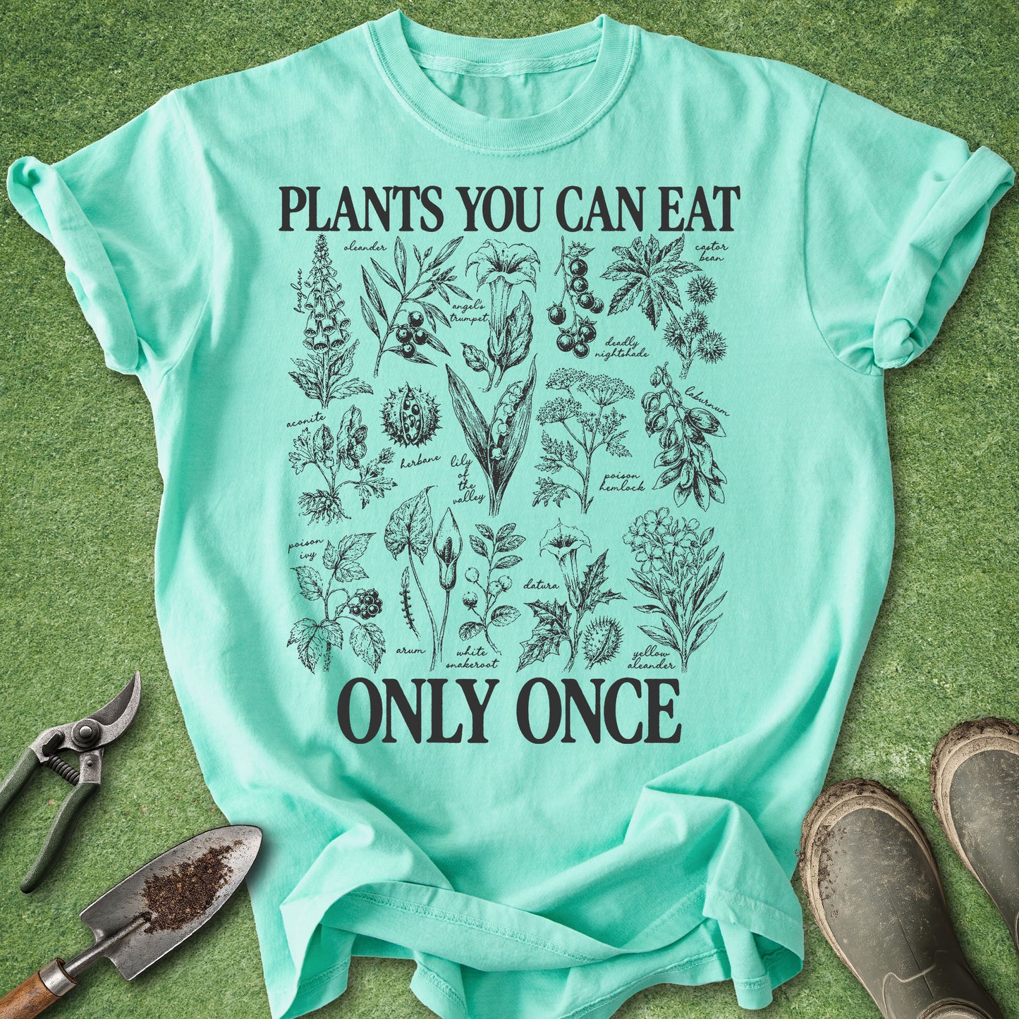 A mint green graphic t-shirt laid flat on a green grass-like surface, surrounded by gardening props including pruning shears, a small hand trowel, and work boots. The t-shirt features a black botanical illustration of edible plants with the text 'PLANTS YOU CAN EAT ONLY ONCE'.