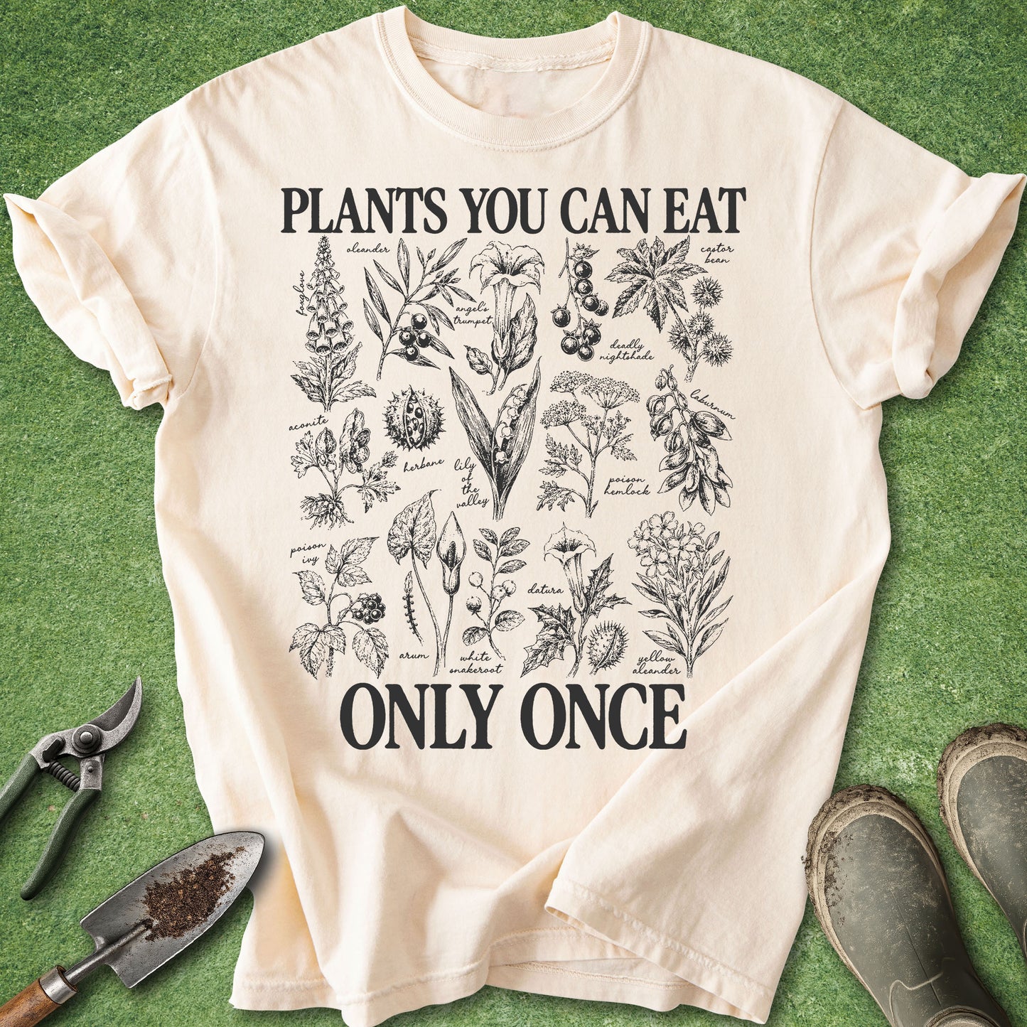A cream-colored graphic t-shirt laid flat on a green grass-like surface, surrounded by gardening props including pruning shears, a trowel, and boots. The shirt features black text reading 'PLANTS YOU CAN EAT ONLY ONCE' with a detailed botanical illustration in the center.