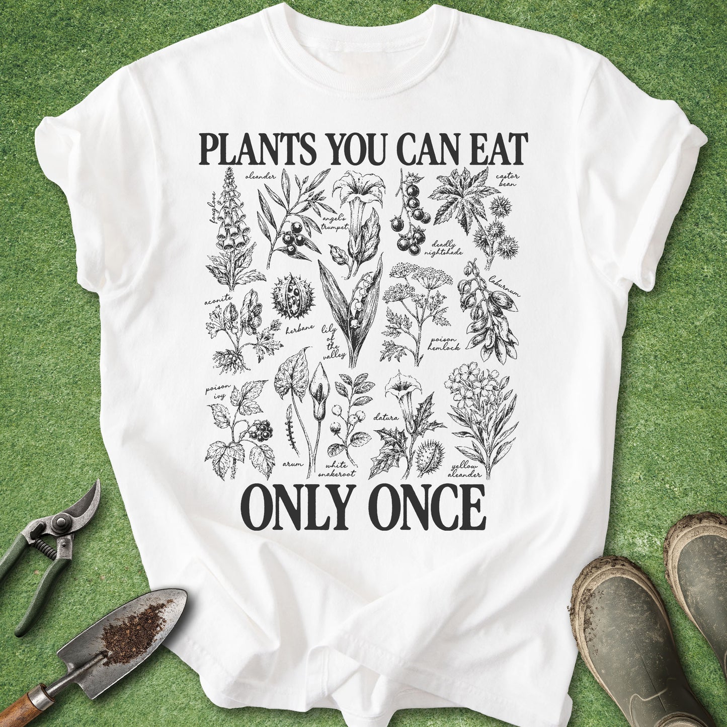 A white graphic t-shirt laid flat on green grass, featuring black botanical illustrations and the text 'PLANTS YOU CAN EAT ONLY ONCE'. Gardening tools and muddy boots are placed around the shirt as props.