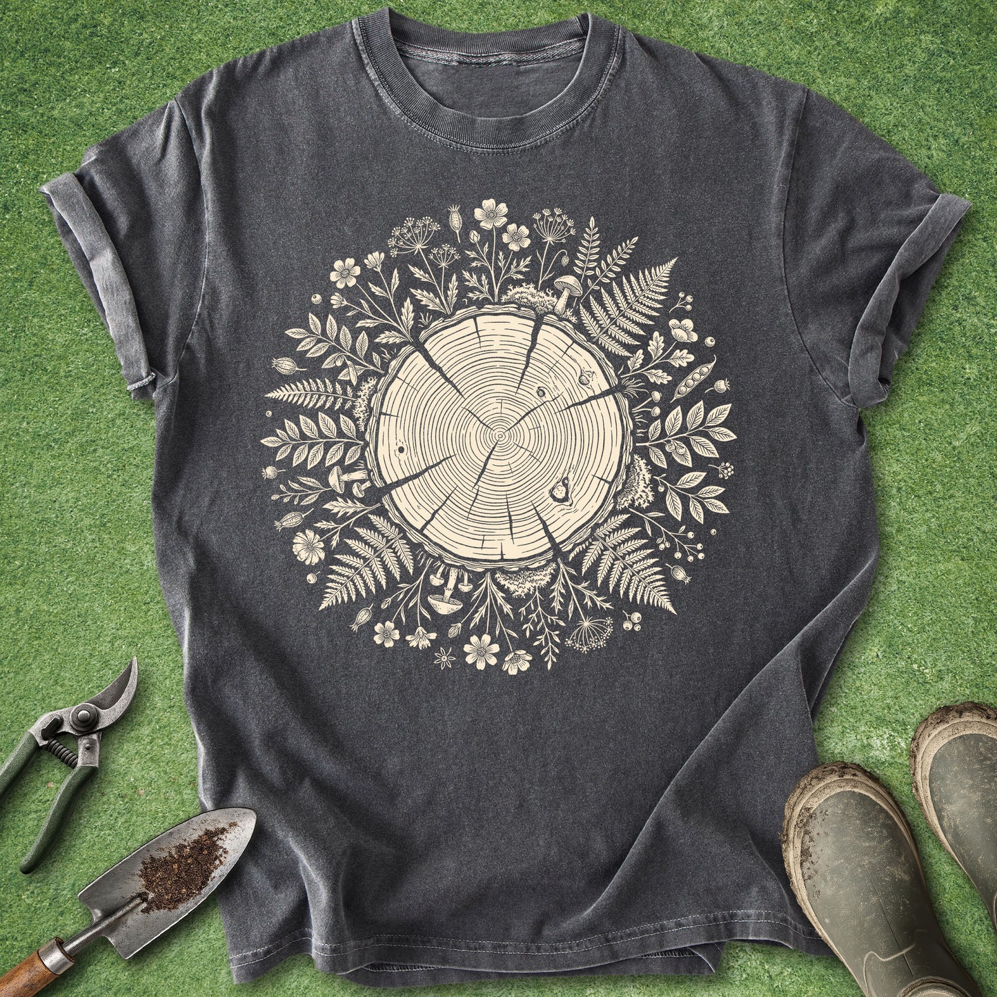 A dark charcoal gray t-shirt with a circular tree stump and floral graphic in cream, laid flat on green grass with gardening tools and boots nearby.