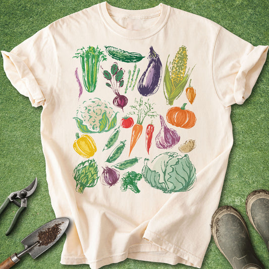 Ivory t-shirt with colorful hand-drawn vegetable illustrations including carrots, cabbage, corn, eggplant, and tomatoes arranged in a garden-inspired design