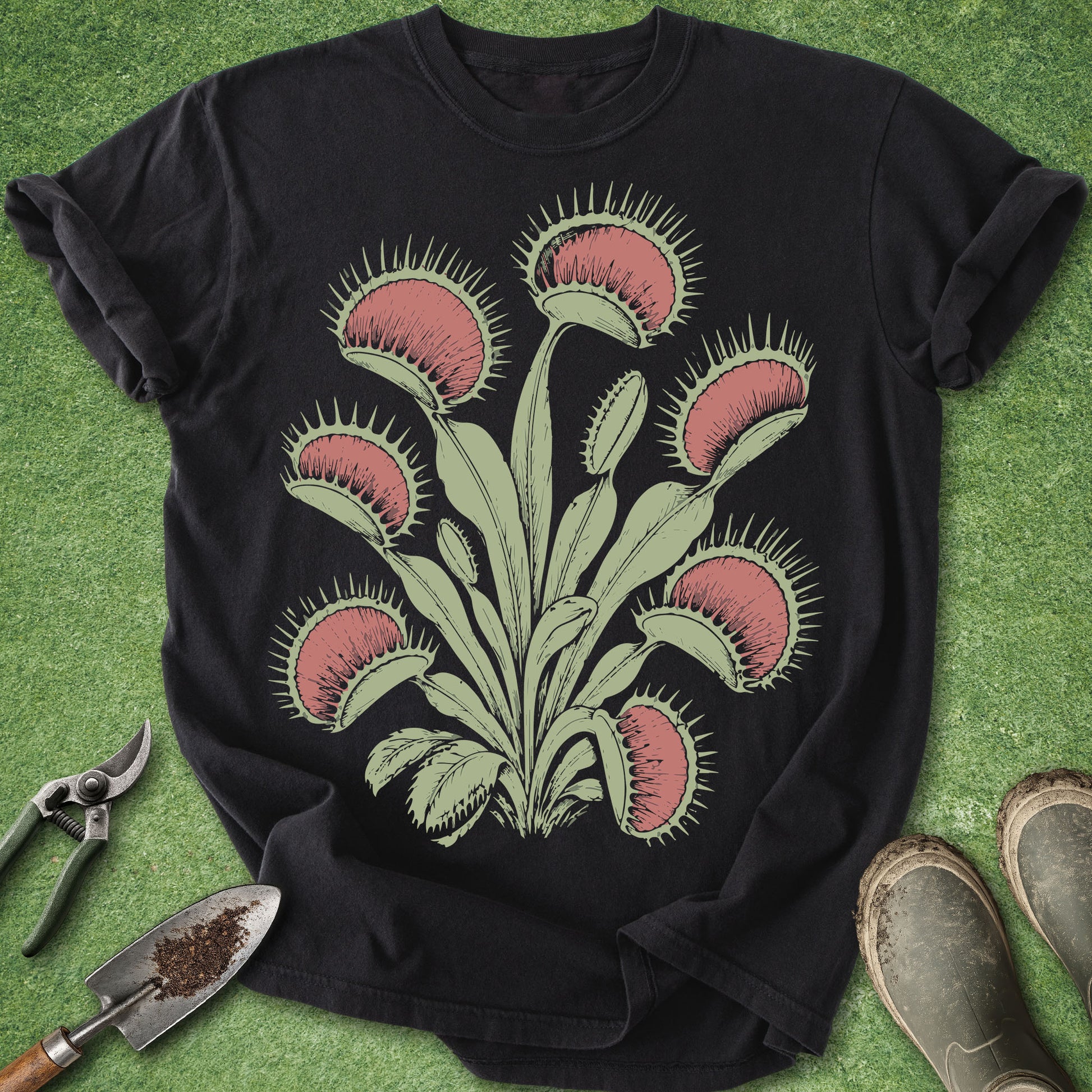 Black t-shirt with Venus flytrap design on a green surface with gardening tools.