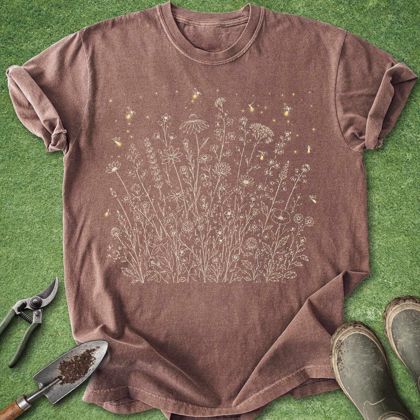 A brown short-sleeve crewneck t-shirt with a white line-art graphic of wildflowers and small yellow butterflies, laid flat on green grass with gardening tools and boots nearby.