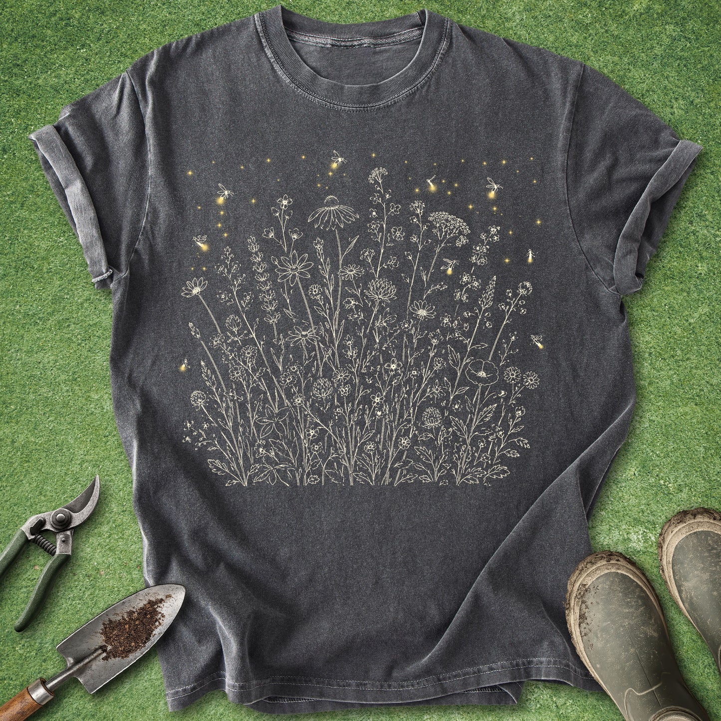A dark charcoal gray t-shirt with a graphic of wildflowers and small yellow insects, laid flat on green grass with gardening tools and boots nearby.