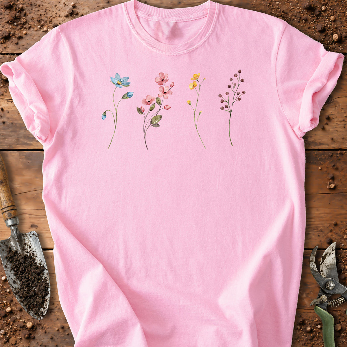 A light pink short-sleeve t-shirt with a watercolor-style floral print on the chest, laid flat on a rustic wooden surface with gardening tools nearby.