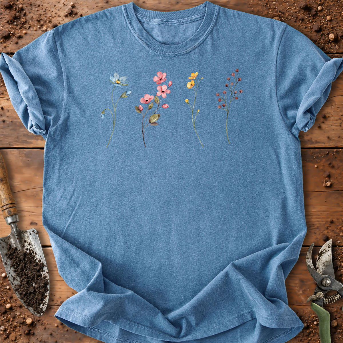 A light blue short-sleeve crewneck t-shirt with a colorful watercolor-style floral print across the chest, styled flat on a rustic wooden surface with gardening tools.