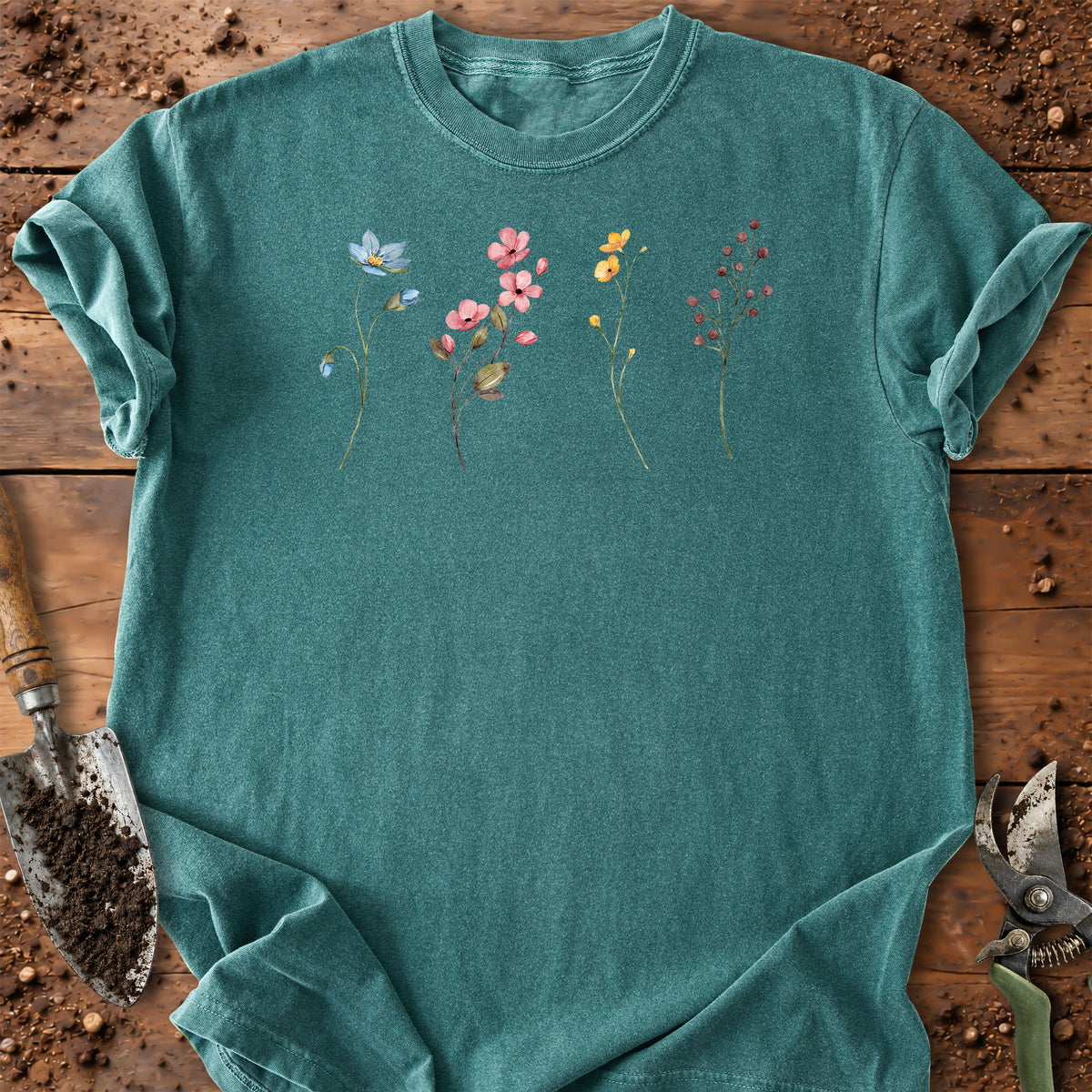 A teal short-sleeve crewneck t-shirt with a delicate watercolor floral print on the chest, laid flat on a rustic wooden surface with gardening tools.