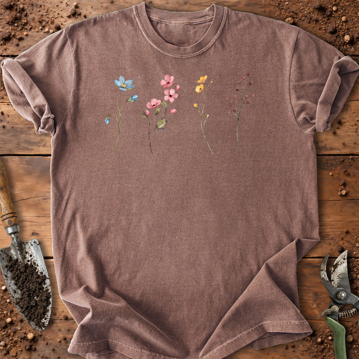 A washed brown short-sleeve t-shirt with a delicate watercolor floral and butterfly print in pink, yellow, and blue across the chest, styled flat on a rustic wooden surface with gardening tools.