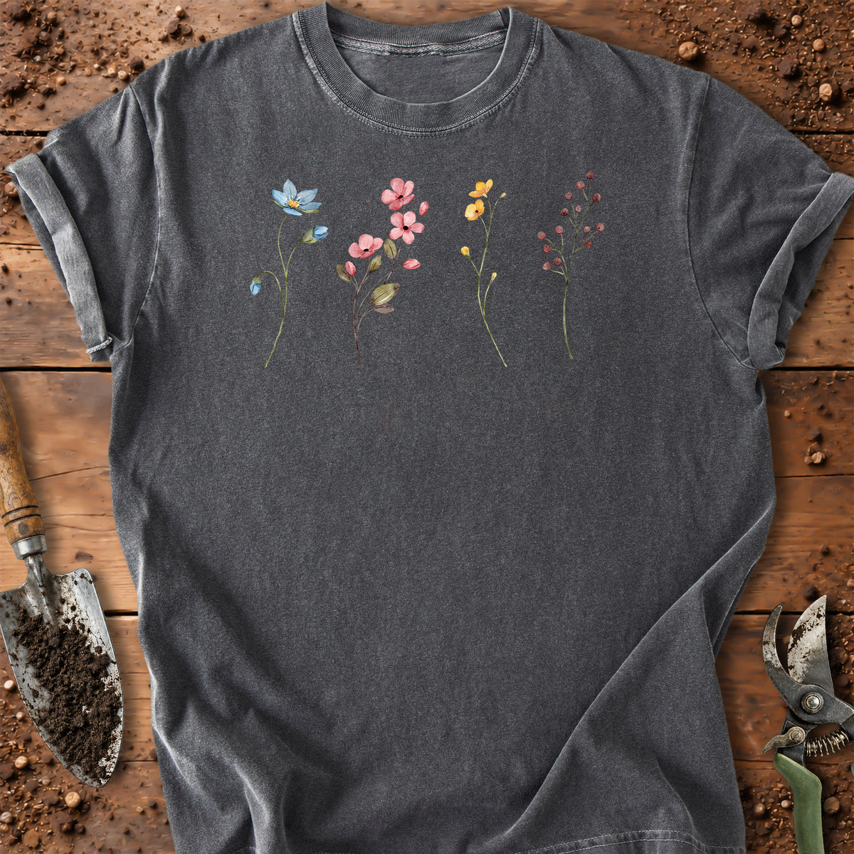 A dark gray t-shirt with a colorful floral graphic, laid flat on a rustic wooden surface with gardening tools like a trowel and pruning shears.
