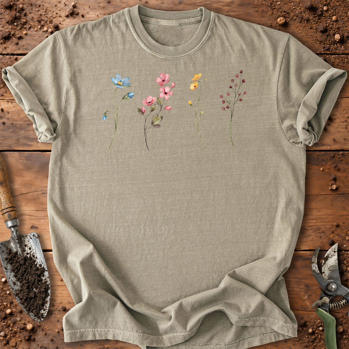 A light beige crewneck t-shirt with a watercolor-style floral print of wildflowers, laid flat on a rustic wooden surface with gardening tools.