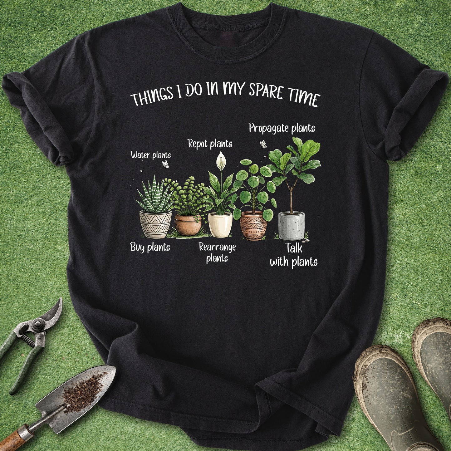 A black graphic t-shirt laid flat on green grass with gardening tools and boots around it. The shirt features a white text graphic reading 'THINGS I DO IN MY SPARE TIME' above an illustrated arrangement of potted plants and labels.