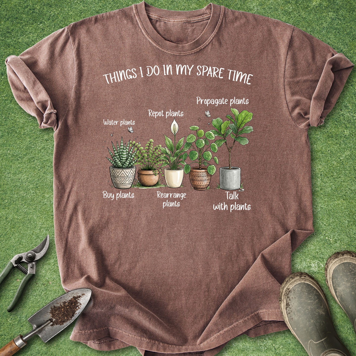 A brown short-sleeve graphic T-shirt laid flat on green grass-like background, with gardening tools and boots around it. The shirt has a printed illustration of potted plants and the text 'THINGS I DO IN MY SPARE TIME' plus labels such as 'water plants,' 'repot plants,' 'propagate plants,' 'buy plants,' 'rearrange plants,' and 'talk with plants.
