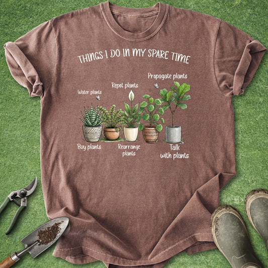 A brown short-sleeve graphic T-shirt laid flat on green grass-like background, with gardening tools and boots around it. The shirt has a printed illustration of potted plants and the text 'THINGS I DO IN MY SPARE TIME' plus labels such as 'water plants,' 'repot plants,' 'propagate plants,' 'buy plants,' 'rearrange plants,' and 'talk with plants.