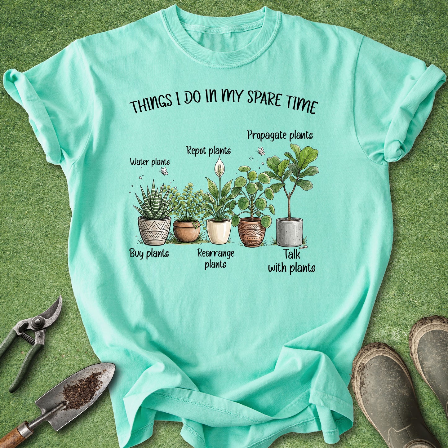 A mint green graphic t-shirt laid flat on a green grass-like surface, with gardening tools and boots around it. The shirt features a plant-care illustration and the text 'THINGS I DO IN MY SPARE TIME.