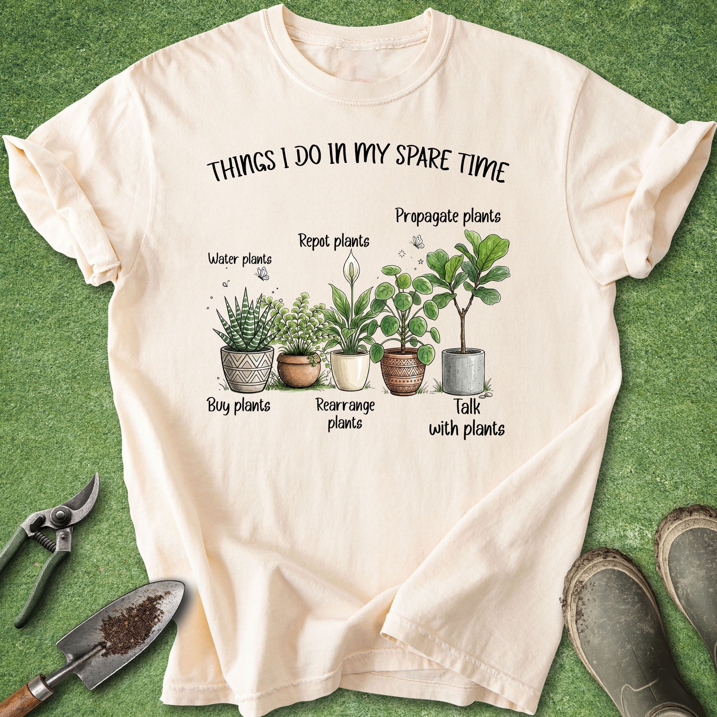 A cream-colored graphic t-shirt laid flat on a green grass-like surface, surrounded by gardening props including pruning shears, a trowel, and boots. The t-shirt features a watercolor-style illustration of various potted houseplants with humorous labels: 'Water plants', 'Repot plants', 'Propagate plants', 'Buy plants', 'Rearrange plants', and 'Talk with plants'.