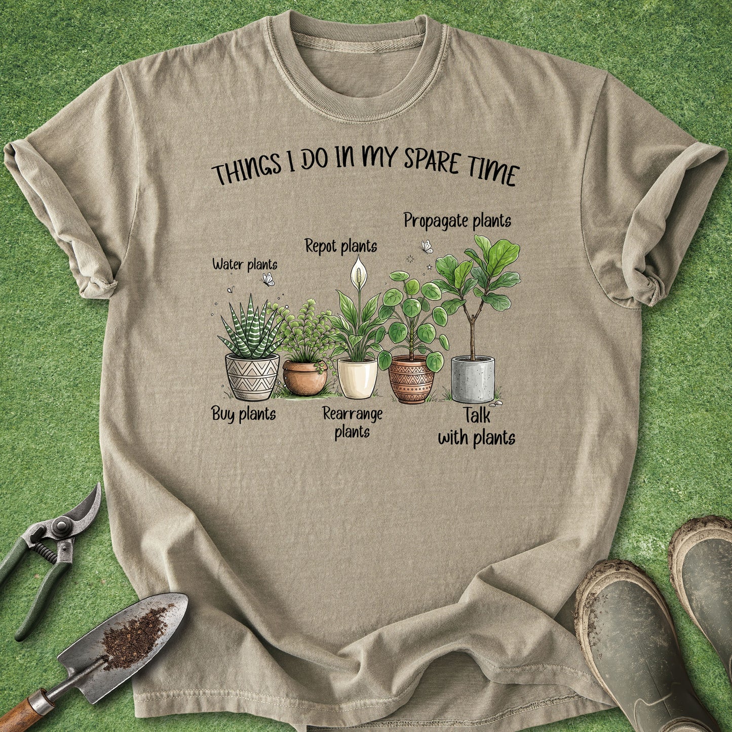 A beige graphic t-shirt laid flat on a green grass-like surface, surrounded by gardening props including pruning shears, a trowel, and boots. The shirt features a printed illustration of potted plants with labels reading 'THINGS I DO IN MY SPARE TIME' and phrases like 'Water plants,' 'Repot plants,' 'Propagate plants,' 'Buy plants,' 'Rearrange plants,' and 'Talk with plants.