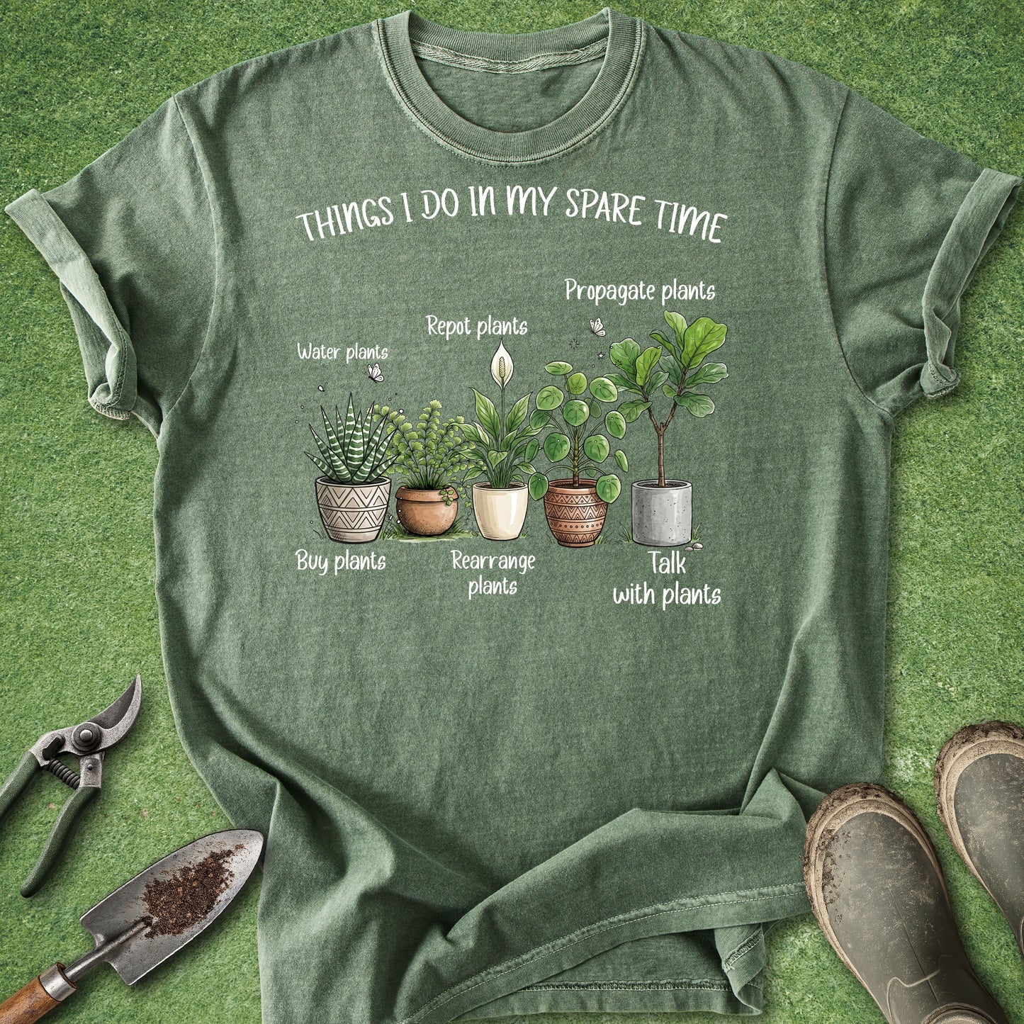 An olive green graphic t-shirt laid flat on a green grass-like surface, surrounded by gardening props including pruning shears, a trowel, and boots. The shirt features a white text graphic reading 'THINGS I DO IN MY SPARE TIME' above an illustration of five potted plants with labels.