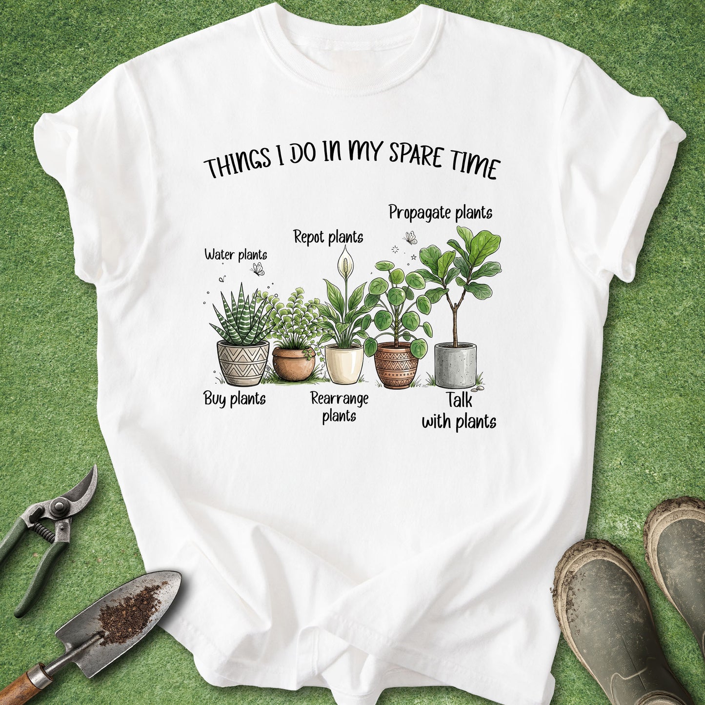 A white graphic t-shirt laid flat on green grass-like background, with gardening-themed printed text and illustrations of potted plants. Surrounding props include pruning shears, a trowel, and gardening boots.