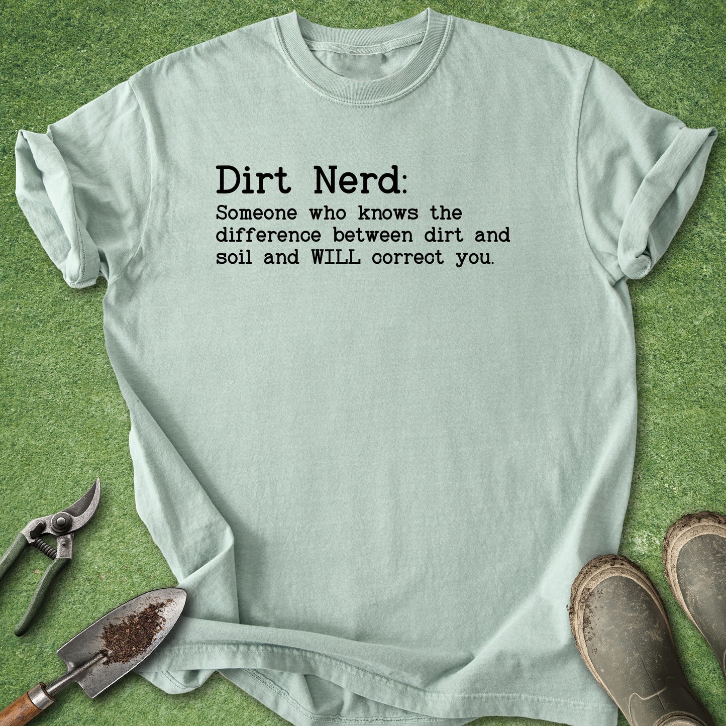 A light sage green graphic t-shirt laid flat on a green grass-like surface, styled with gardening tools (trowel, pruning shears) and muddy boots. The t-shirt features black printed text that reads: 'Dirt Nerd: Someone who knows the difference between dirt and soil and WILL correct you.